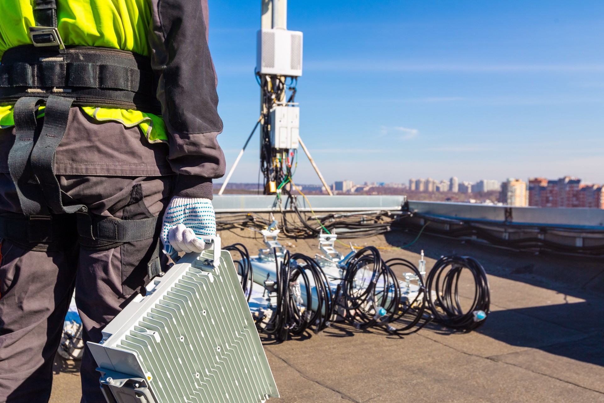 Professional industrial climber in uniform with telecomunication equipment in his hand and antennas of GSM DCS UMTS LTE bands, outdoor radio units on the roof. Working process of upgrading or swap telecommunication equipment.